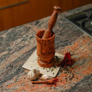 May include: A wooden mortar and pestle set with a rich, reddish-brown grain pattern. The mortar is cylindrical, and the pestle is tapered. The set is accompanied by garlic cloves, red chili peppers, and spices, suggesting culinary use.
