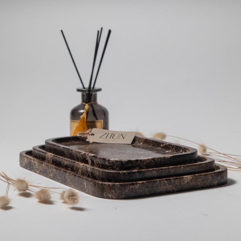 May include: A set of three rectangular brown marble trays, stacked on top of each other. A dark glass diffuser bottle with black reeds and a tag that reads "ZIJUN" is in the background. The trays are a decorative home decor item.