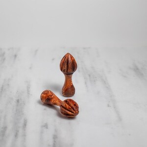 May include: Two wooden citrus reamers. One is upright, the other is lying down. The reamers are made of a reddish-brown wood with a ribbed design on the reaming head. The reamers are designed for juicing citrus fruits.