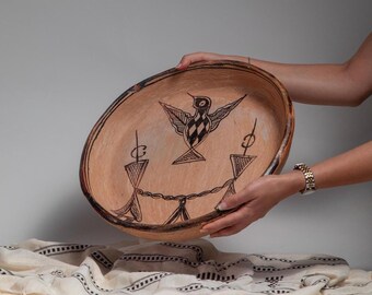 May include: A large, oval-shaped, hand-painted ceramic bowl with a bird and geometric designs in black. The bowl is held by two hands, and the background is a neutral color.