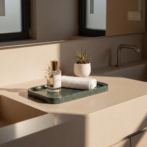 May include: A bathroom countertop arrangement featuring a dark green marble tray holding a perfume bottle, a rolled white towel, and a small potted plant. The perfume bottle has a gold cap. The countertop is a light beige color.