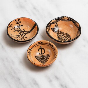 May include: Three small, hand-painted ceramic bowls with a terracotta base and black detailing. Each bowl features a unique design: a branch, a fish, and a geometric symbol. The bowls have a black rim and are arranged on a white marble surface.