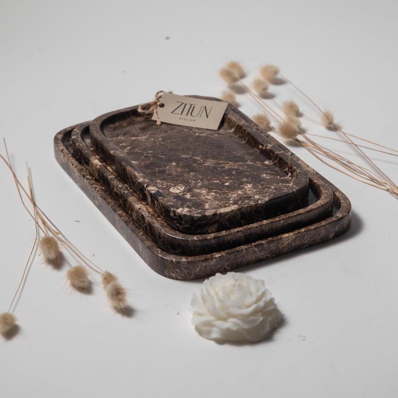 May include: Set of three rectangular decorative trays in varying sizes. The trays have a marbled brown and cream pattern. A small tag with the text "ZIJUN" is attached to the top tray. The trays are arranged on a white surface with dried flowers and a white flower.
