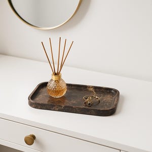 May include: A decorative tray with a marble-like pattern holds a glass diffuser bottle with reeds and a gold-colored ring. The diffuser bottle has a textured, round shape. The tray is rectangular with rounded corners.