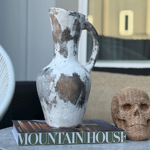 May include: A white and brown decorative jug with a handle, on a table with a book that says "MOUNTAIN HOUSE" and a woven skull. The jug has a rustic, aged appearance.