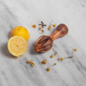May include: A wooden citrus reamer with a carved design, next to a halved lemon and a whole lemon. Dried chamomile flowers and spices are scattered around the reamer and lemons. The reamer is made of a dark wood.