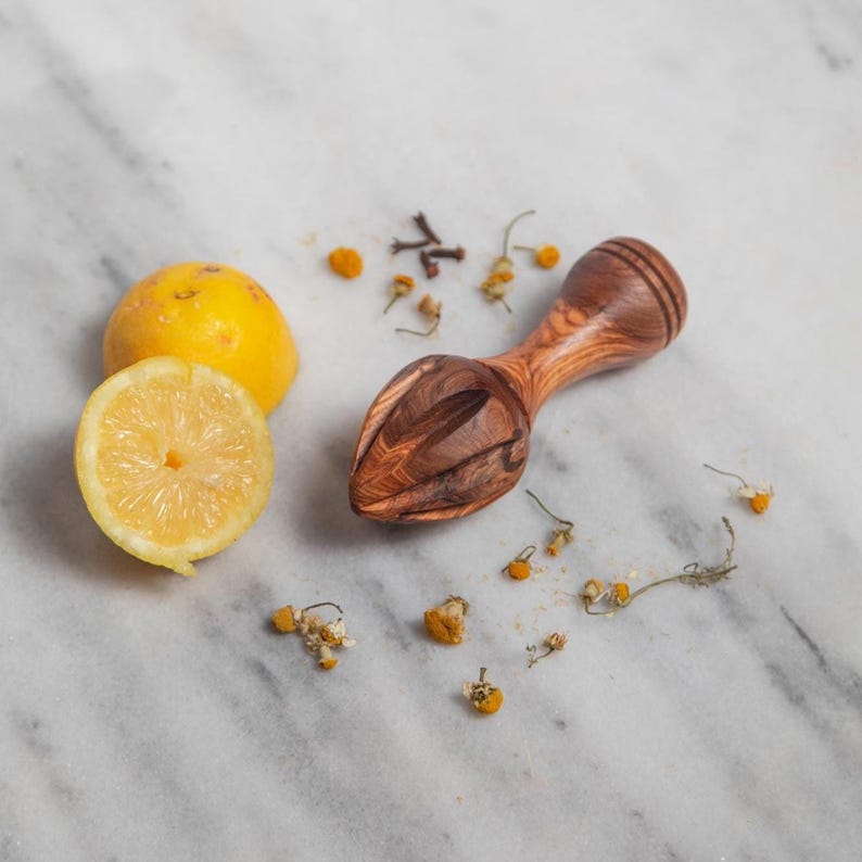 May include: A wooden citrus reamer with a carved design, next to a halved lemon and a whole lemon. Dried chamomile flowers and spices are scattered around the reamer and lemons. The reamer is made of a dark wood.