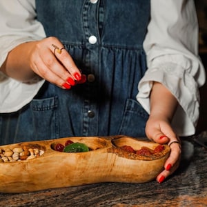May include: A wooden serving bowl with three compartments, filled with pistachios, kiwi, and dried fruit. The bowl is held by a person wearing a denim dress and white shirt. The person is adding red berries to the bowl.