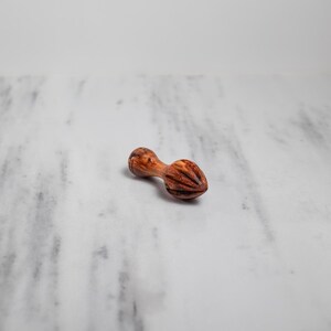 May include: A wooden citrus reamer with a double-ended design. The reamer is made of a light brown wood with darker brown grain patterns. The reamer is lying on a white and gray marble surface.