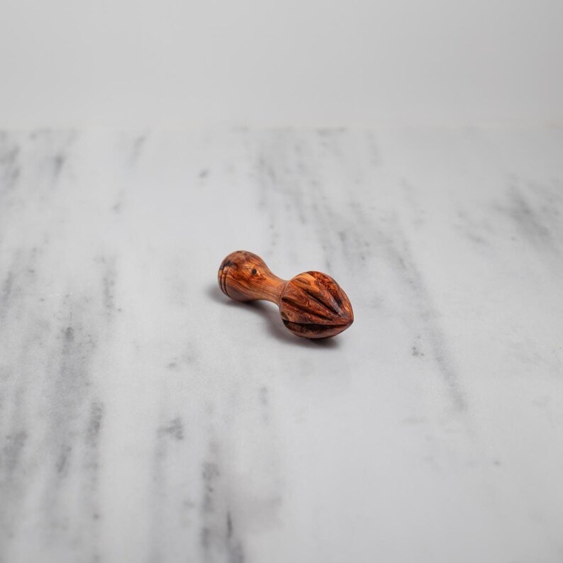 May include: A wooden citrus reamer with a double-ended design. The reamer is made of a light brown wood with darker brown grain patterns. The reamer is lying on a white and gray marble surface.