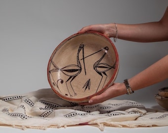 May include: A beige ceramic bowl with a red rim, featuring a black illustration of two birds. The bowl is held in front of a white background. The bowl is part of a collection of handmade pottery.
