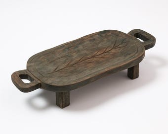 May include: A rustic, rectangular wooden serving tray with a dark, weathered finish. The tray features a carved leaf design and two handles. It stands on four short, square legs, offering a raised presentation for food or decor.