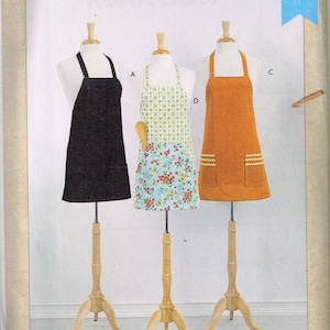 May include: Three unisex aprons on display stands. The aprons are black, orange, and white with a floral pattern. The pattern is a small floral print on a light blue background. The aprons are labelled A, C, and D. The text "Kwik Sew K4287" is in the top right corner of the image. The text "Unisex Aprons" is at the bottom of the image.