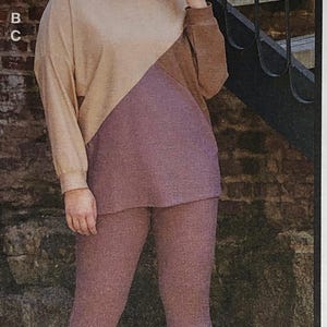 May include: A two-toned hooded sweatshirt with a beige top and a mauve bottom. The sweatshirt has a relaxed fit and a drawstring hood. The model is wearing the sweatshirt with mauve leggings and brown sneakers.