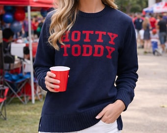 Może przedstawiać: Granatowy sweter z dzianiny z długim rękawem z czerwonym napisem HOTTY TODDY na piersi, zestawiony z białymi szortami dżinsowymi z postrzępionym brzegiem. Sweter ma prążkowany okrągły dekolt, mankiety i dół z małymi rozcięciami po bokach.