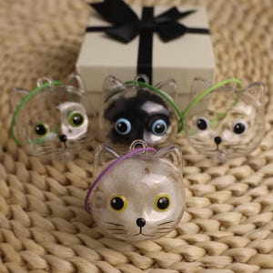 Puede incluir: Cuatro adornos transparentes con forma de gato con caras pintadas y lazos de cuerda de colores. Un adorno tiene una cara de gato negro, mientras que los otros tienen patrones atigrados. Una caja de regalo con un lazo negro está en el fondo.