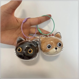 Puede incluir: Dos llaveros con forma de gato. Uno es negro con ojos amarillos, el otro es marrón con pelo castaño. Ambos están encerrados en plástico transparente con un lazo de metal y un cable de alambre de color.