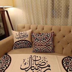May include: Beige sofa with two decorative throw pillows and a table runner. The pillows and runner feature black Arabic calligraphy and burgundy, green, and cream geometric patterns. A floor lamp is visible in the background.