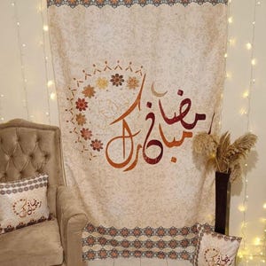 May include: Beige tapestry with ornate floral and geometric patterns, featuring Arabic calligraphy in shades of orange and brown. Decorative pillows and a chair are in the scene. The tapestry is decorated with a floral design and a crescent moon.