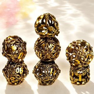 May include: A set of five spherical, gold-colored metal dice featuring an intricate, openwork design of intertwined vines and leaves. Each die is hollow with cut-out numbers integrated into the filigree pattern, creating a textured, cage-like appearance.