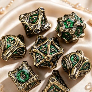 May include: A set of seven polyhedral dice featuring an antique gold-toned metal finish with intricate, branch-like raised textures and recessed emerald green enamel inlays. Each die displays clear, dark-colored numbers on its various faces.
