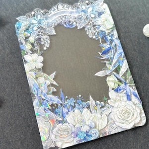 Pode incluir: Uma moldura de plástico transparente com um design floral. A moldura é decorada com flores brancas e azuis e tem um acabamento holográfico.