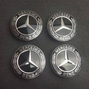Puede incluir: Cuatro tapacubos Mercedes-Benz. Cada tapacubo presenta un centro negro con el logotipo plateado de Mercedes-Benz y la palabra "BENZ" debajo. Los tapacubos están rodeados por un diseño de corona plateada.
