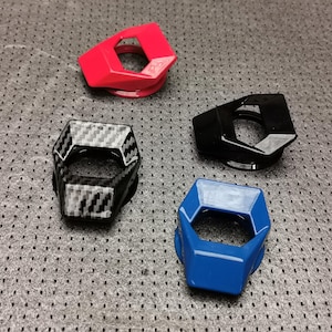 Op de afbeelding: Vier zeshoekige auto-accessoires in rood, zwart, koolstofvezel en blauw. Elk heeft een centrale opening en een geometrisch ontwerp. Het koolstofvezelstuk heeft een textuurpatroon. Ze liggen op een grijze, gestippelde ondergrond.