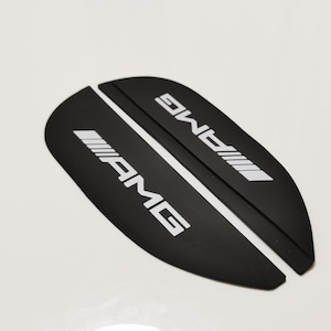 Może przedstawiać: Dwie czarne, zakrzywione osłony progów drzwi samochodowych z białym tekstem i logo "AMG". Ochraniacze wykonane są z matowego materiału i są przeznaczone do montażu na progach drzwi pojazdu.