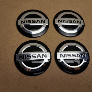 May include: Four black and silver Nissan wheel center caps. Each cap features the Nissan logo in silver lettering on a silver background, surrounded by a black outer ring. The caps are circular and appear to be new.