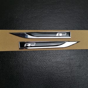 Pode incluir: Dois emblemas de carro R Line pretos e prateados. Cada emblema tem uma forma longa e elegante com uma extremidade pontiaguda. O logótipo R Line é branco. Os emblemas estão numa superfície de cartão castanho.