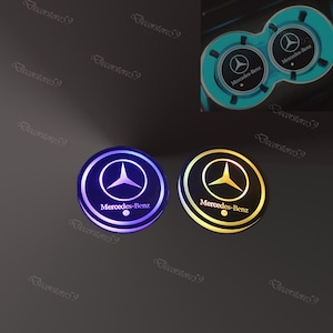 May include: Two illuminated Mercedes-Benz car cup holder inserts. One glows purple, the other gold, both with the Mercedes-Benz logo and text. The inserts are round and have a sleek, modern design.