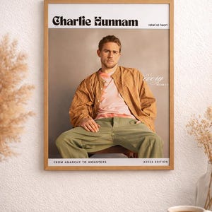May include: A framed poster featuring Charlie Hunnam. He is wearing a pink shirt, a brown striped jacket, and green pants. The poster includes the text "Charlie Hunnam" and "rebel at heart". Additional text reads "FROM ANARCHY TO MONSTERS" and "#2026 EDITION".