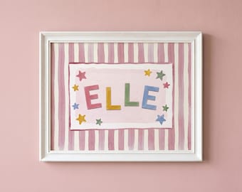 May include: A framed print featuring the name ELLE in colourful block letters centred on a light pink background. The design includes small multi-coloured stars surrounding the name, all set against a backdrop of vertical pink and white stripes.