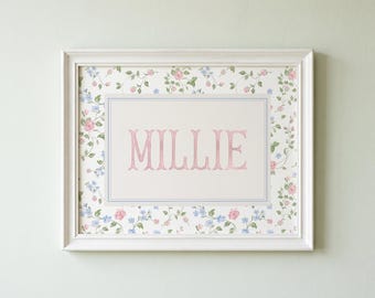 May include: A rectangular white frame containing a print with the name MILLIE in pink capital letters. The background features a floral pattern with small pink roses and blue flowers on a white field, bordered by a thin blue line rectangle.