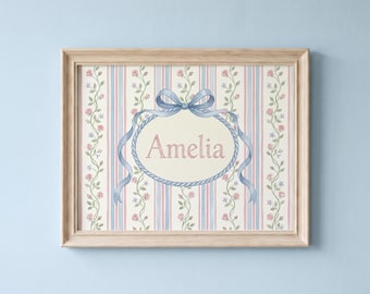 May include: A rectangular wooden frame containing a print with a vertical striped background in blue, pink, and cream. The print features climbing rose vines, a central oval rope border, a blue ribbon bow, and the name Amelia in pink lettering.