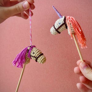 Könnte beinhalten: Zwei handgefertigte Häkel-Einhornköpfe auf Holzstäben. Einer hat eine lila Mähne und der andere eine orangefarbene Mähne. Beide haben braune Zaumzeugdetails und ein Horn. Der Hintergrund ist einfarbig rosa.