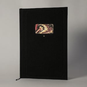 May include: A black hardcover book with a decorative rectangular inset featuring a swirling pattern of red, brown, and cream colors. A small star is below the inset. A black ribbon bookmark is visible.