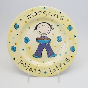 May include: A yellow ceramic plate with a cartoon illustration of a person holding a plate of potato latkes. The plate is decorated with blue and yellow confetti and the text "morgan's potato latkes".