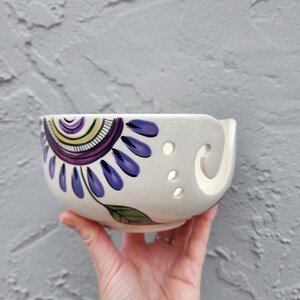 May include: White ceramic yarn bowl with a purple, blue, and green floral design. The bowl has holes around the rim for yarn to pass through.
