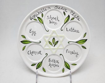 Op de afbeelding: Een wit keramiek Seder-bord met een zespuntige ster in het midden en zes secties rond de ster. Elke sectie is gelabeld met een ander item voor het Seder-bord: schenkelbeen, ei, charoset, bittere kruiden, peterselie en sla. Het bord is versierd met groene bladeren rond de rand en de woorden "Annabelle's Seder Plate" staan rond de bovenkant van het bord.