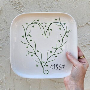 May include: White square ceramic plate with a green leafy vine design forming a heart shape. The number 01867 is written in black cursive on the plate.