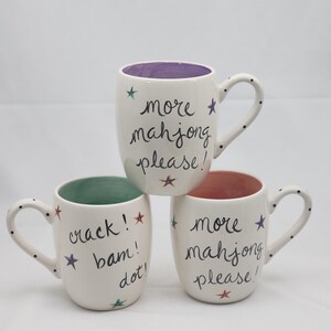May include: Three white ceramic mugs with black polka-dot handles. Two mugs feature the text "more mahjong please!" with colorful stars, one with a purple interior and one with a pink interior. The third mug has a green interior and the text "crack! bam! dot!"