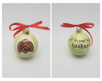 May include: A green Christmas ornament with a painted image of a brown dog and the text "Forever in our hearts Tucker". The ornament is tied with a red ribbon.