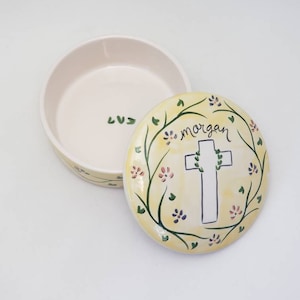 May include: A yellow ceramic trinket box with a white cross and floral design. The lid has the name "morgan" written on it.