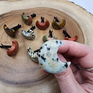May include: A collection of crescent moon shaped car air fresheners made from various types of stone, including dalmatian jasper, tiger's eye, and red jasper. The air fresheners are displayed on a wooden surface.