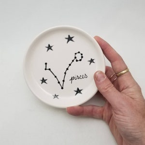 May include: White ceramic dish with a black outline of the Pisces constellation and the word "Pisces" written in black cursive. The dish is decorated with black stars.