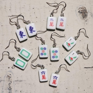 May include: A collection of five pairs of dangle earrings made from white rectangular mahjong tiles. Each tile features distinct colorful symbols, including Chinese characters, floral patterns, and circular designs, suspended from silver-toned metal hooks.