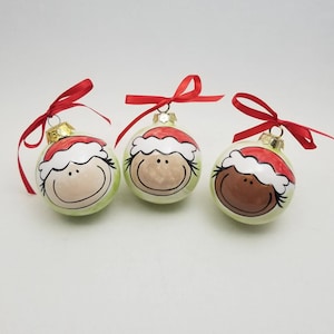 May include: Three green Christmas ornaments with painted faces and red Santa hats. Each ornament has a different skin tone.
