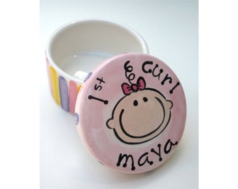 May include: A pink ceramic keepsake box with a colorful striped pattern on the bottom. The lid features a hand-painted illustration of a baby girl with a pink bow and the words "1st girl! maya".